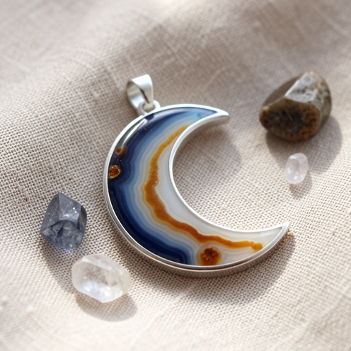 Pendentif en forme de lune serti d’une agate aux nuances bleues, ambrées et blanches, posé sur un tissu en lin beige avec quelques cristaux naturels.
